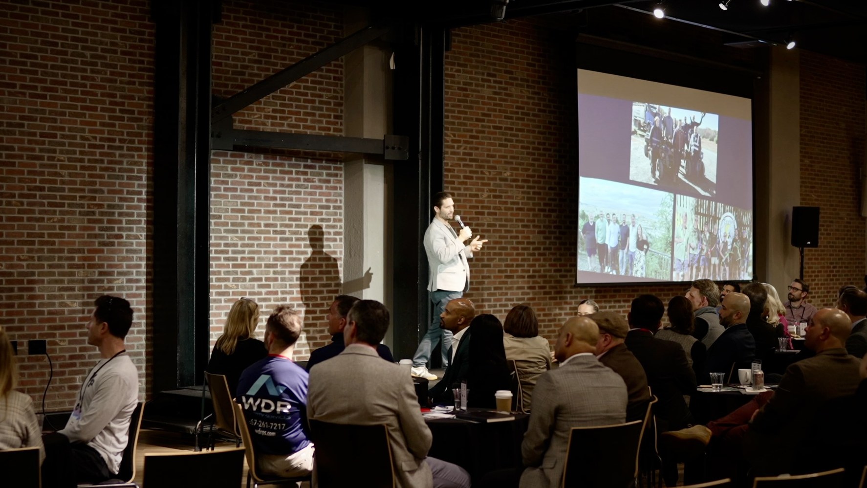 Griff on stage speaking to a room of founders, with travel photos on the screen behind him